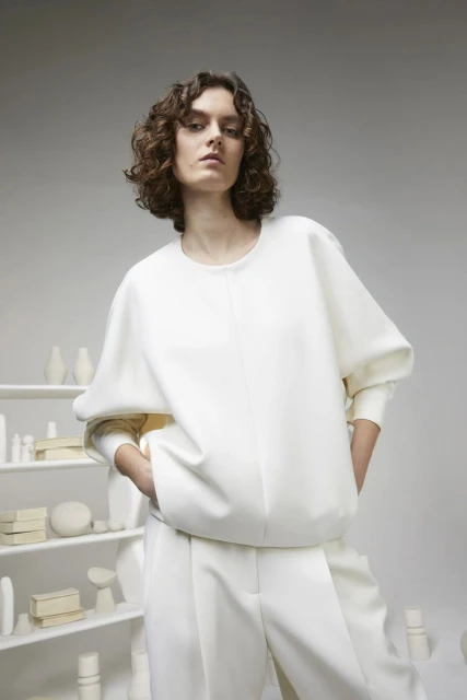 A woman poses in a white oversized sweater and wide-leg trousers in a neutral studio, highlighted by soft, diffused lighting.