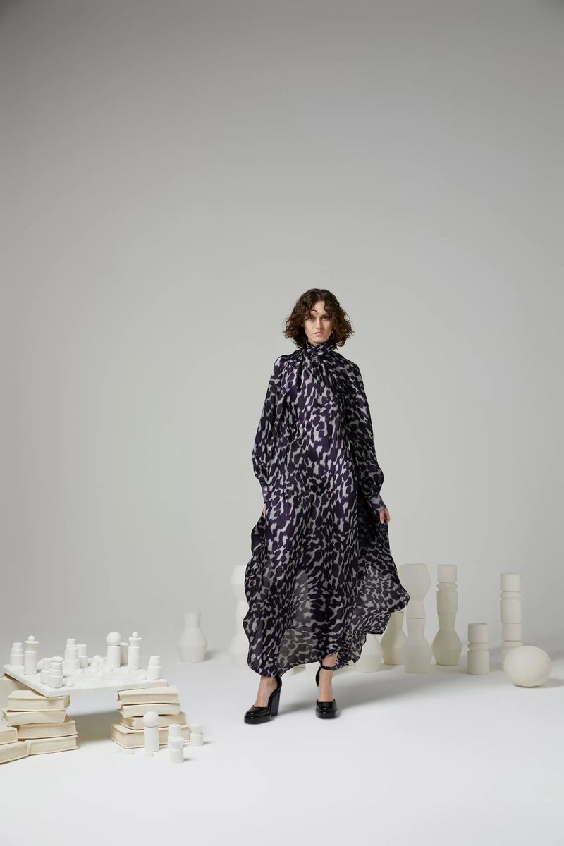 A woman in a flowing black and white patterned dress poses in a white studio surrounded by white chess pieces and ceramic objects.
