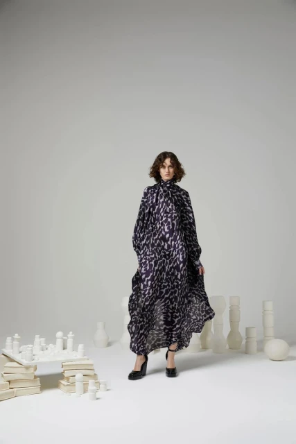 A woman in a flowing black and white patterned dress poses in a white studio surrounded by white chess pieces and ceramic objects.