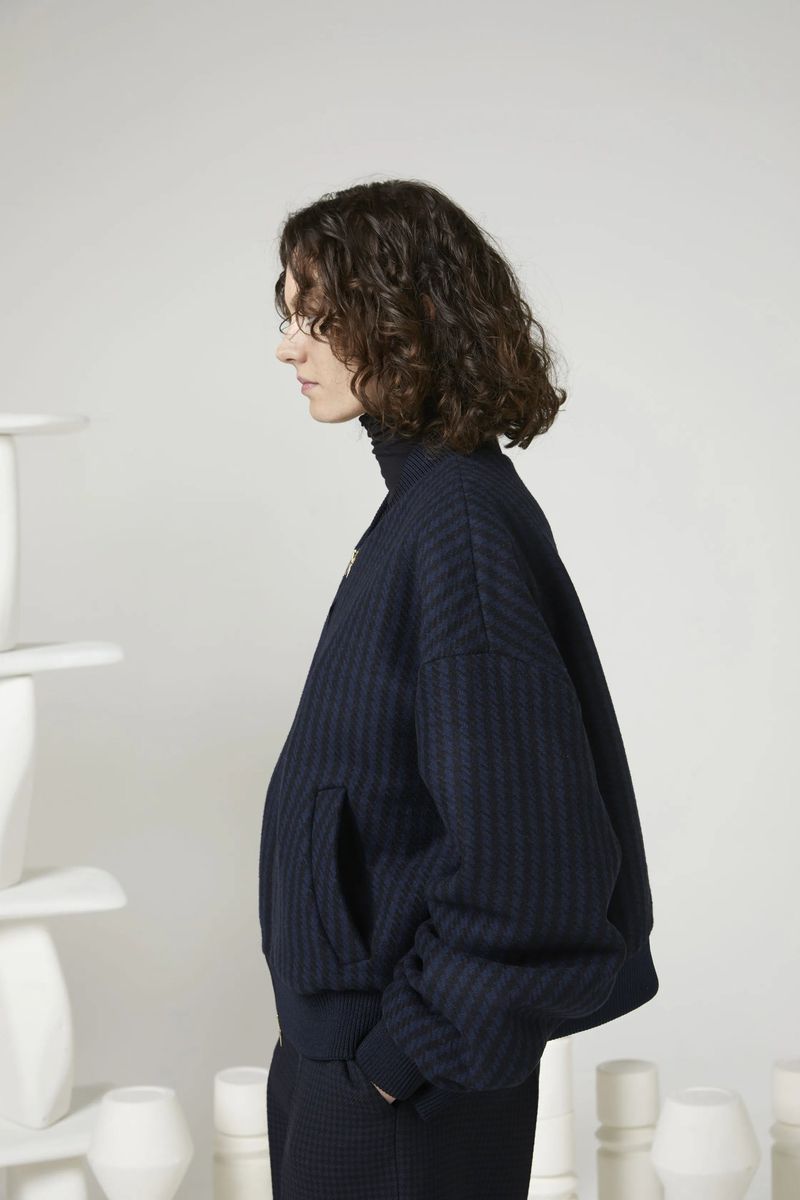 Profile shot of a person with curly brown hair wearing a dark blue textured jacket and matching pants against a gradient white backdrop.