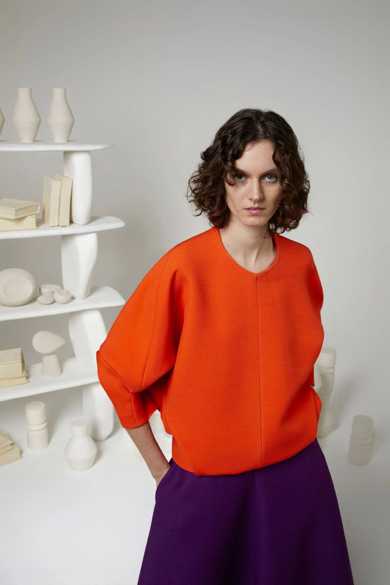 Fashionable woman in a studio setting with orange top and purple skirt poses near a decorative shelf of vases and books.