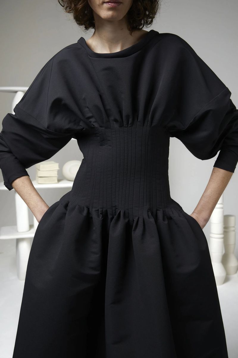 Model wearing a Maison Rabih Kayrouz black dress with a ribbed corset waist and puffed sleeves against a neutral studio set.