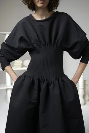 Model wearing a Maison Rabih Kayrouz black dress with a ribbed corset waist and puffed sleeves against a neutral studio set.