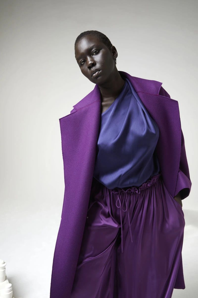 Elegant woman in a purple coat and loose pants poses in a studio with soft, diffused lighting, presenting a fashion editorial shot.