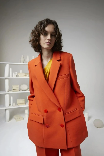 Adult woman in an orange suit poses in a studio with a shelf of decorative white objects in the background. Fashion editorial.