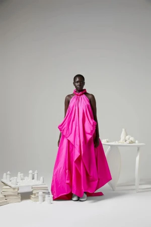 Model standing in a vibrant pink halter neck gown against a minimalist white studio background with sculptural objects