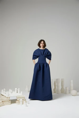Fashion model Hannah Claverie wearing a sculptural navy blue floor-length gown in a studio setting with white chess-like props.