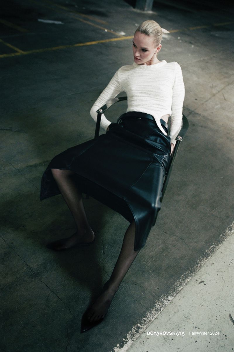 Fashion shot of a blond woman sitting on a chair in a parking garage wearing a white sweater and black skirt, looking off to the side.