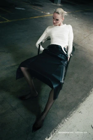 Model with blonde hair seated in a dark chair wearing a white long-sleeve top and black skirt in a concrete garage interior.