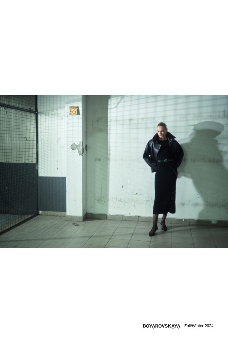 Fashion model poses in a black jacket and skirt in an industrial-style indoor setting with bars and tiled floors for a fashion editorial.