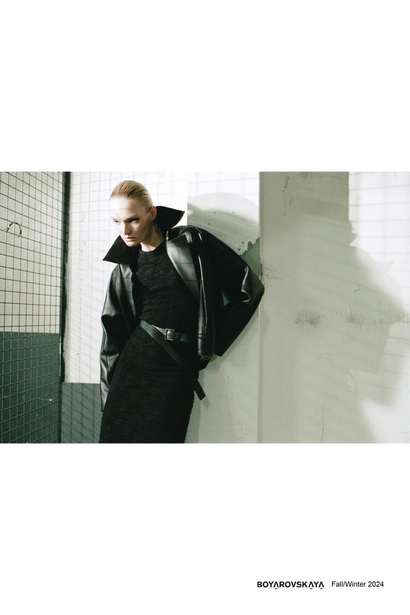 Model wearing a black leather jacket and black dress posing against an industrial concrete wall for Boyarovskaya Fall Winter 2024.