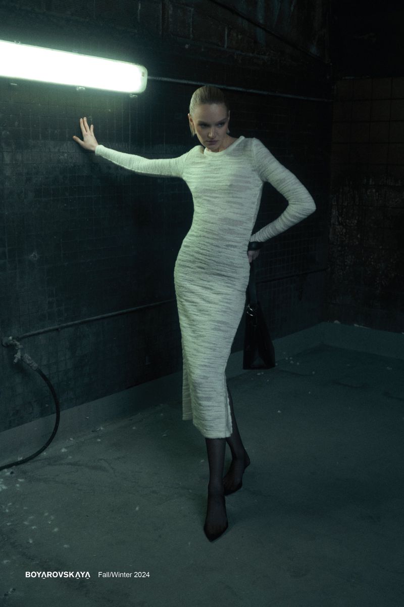Indoor fashion shot of a slim adult woman in a long-sleeved midi dress posing and touching a concrete wall under artificial light.