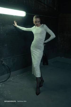 Model wearing a long-sleeved cream textured midi dress leaning against a dark wall under an illuminated tube light.