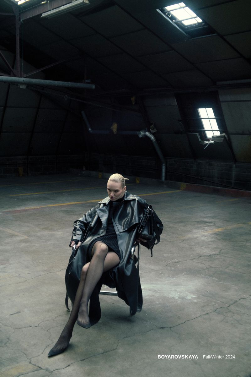 A fair-skinned woman sits in a chair, dressed in black trench coat, stockings, and heels, inside an industrial building with dark lighting.