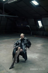 Model wearing an oversized black leather trench coat seated on a chair inside a dark industrial warehouse space.