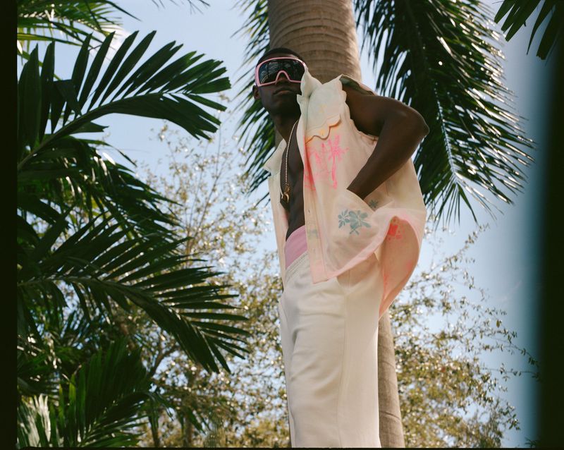 Man in white pants, pink trimmed top, and pink sunglasses standing in front of a palm tree with a blue sky and palm fronds.