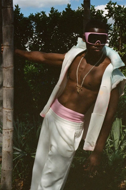 An adult man with an athletic build poses outdoors, shirtless with a white jacket and pants, accented by pink eyewear and waistband.