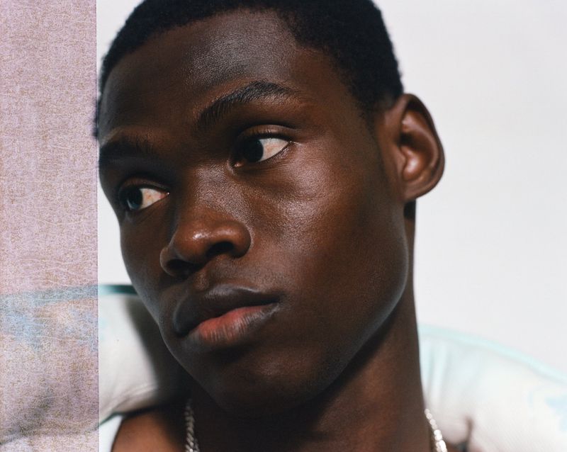 Close-up portrait of a young Black man with dark hair and a silver chain, looking slightly off-camera against a light background.