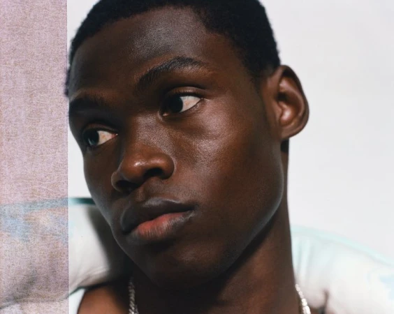 Close-up portrait of a young Black man with dark hair and a silver chain, looking slightly off-camera against a light background.