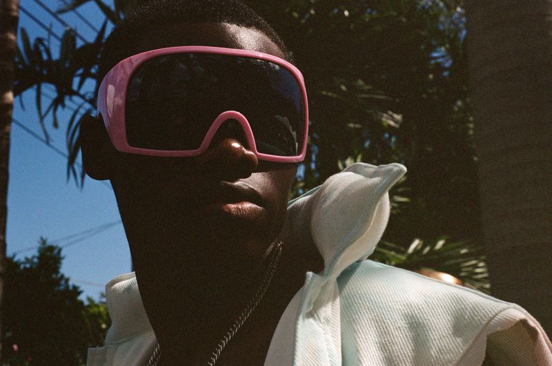 Close-up shot of a stylish Black adult male model wearing large pink sunglasses and a light-blue denim jacket outdoors in sunny weather.