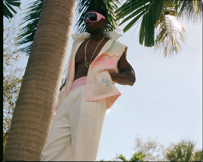 Stylish adult male poses outdoors in a sleeveless top and white pants with pink trim, set against a background of palm trees and sky.