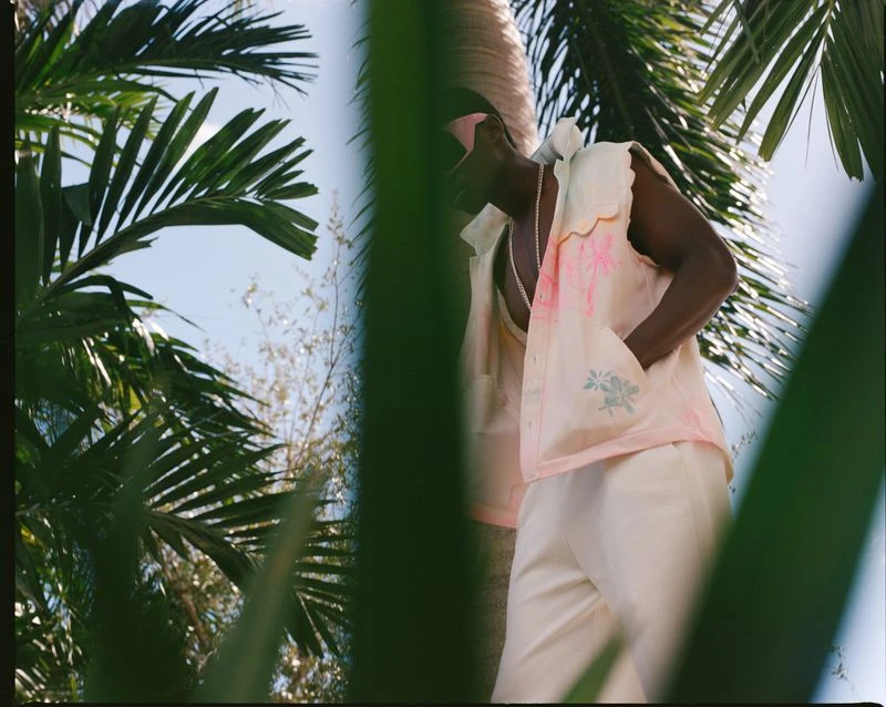 Person in linen suit with floral vest surrounded by palm leaves on a sunny day in tropical outdoor environment.