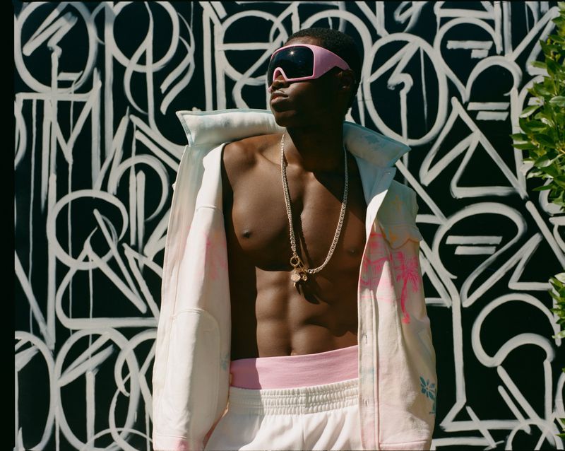 Man with bare chest wearing pink visor and white jacket stands against a black and white painted wall outdoors in daylight.