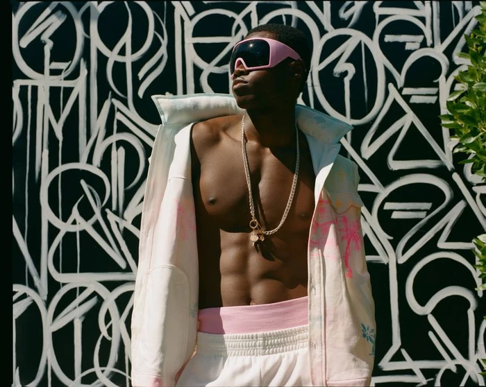 Man wearing pink visor sunglasses, a white jacket, and layered chains against a black and white graffiti wall backdrop.