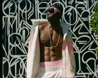 Man wearing pink visor sunglasses, a white jacket, and layered chains against a black and white graffiti wall backdrop.