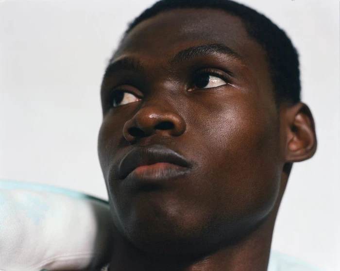 Close-up portrait of a Black man looking upward against a plain light background with fabric draped on his shoulder.