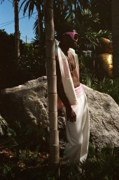 Man wearing pink sunglasses and white trousers leaning against a palm tree outdoors near a large rock for Zara Miami Capsule Collection