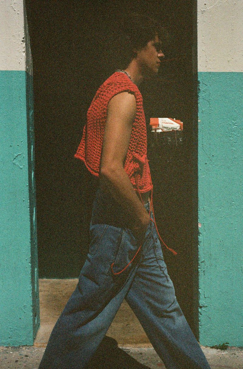 Young adult male in a red knit sleeveless top walks past an aqua and white wall. Fashion shoot with cool, casual style and good light.