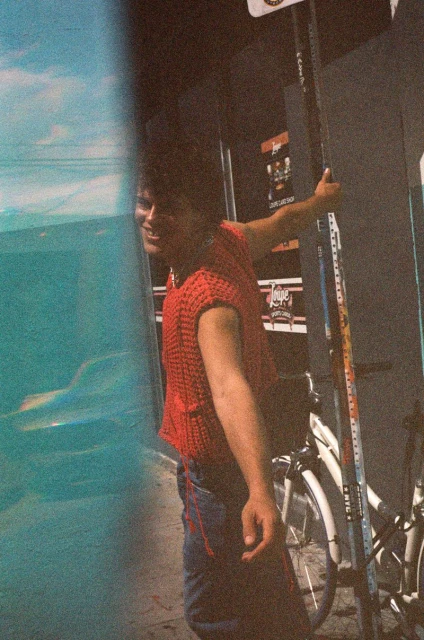 An adult man in a red knit top stands on an urban street next to a bicycle and a bright blue mural with a smile.