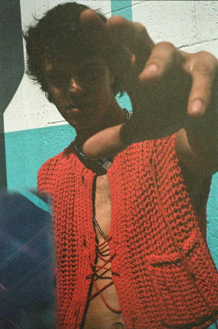A young adult poses wearing a red knit jacket and jewelry, pointing at the camera against a painted wall backdrop. Fashion editorial.
