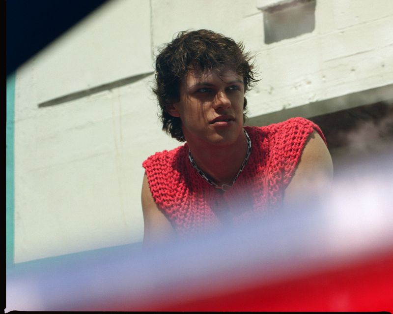 An adult male poses in front of a white building with a red foreground, wearing a red, knitted shirt and a silver chain necklace.