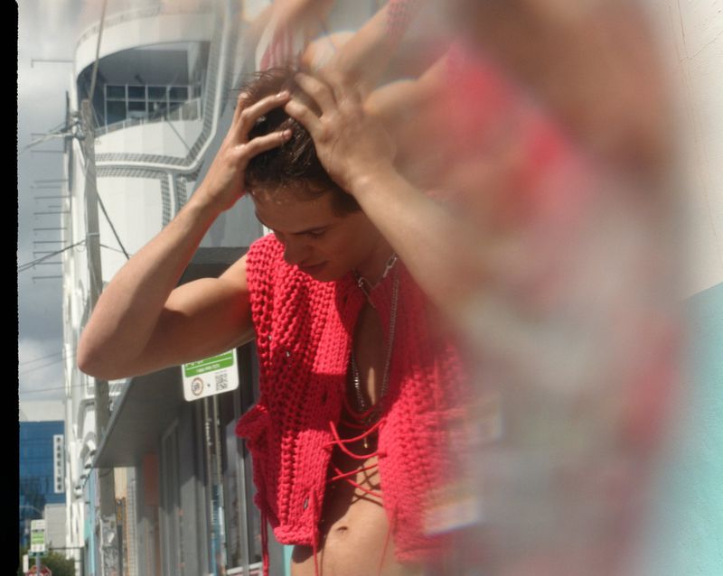 Adult in red knitwear poses with hands on head outdoors, captured in bright, diffused daylight in a casual fashion editorial style.