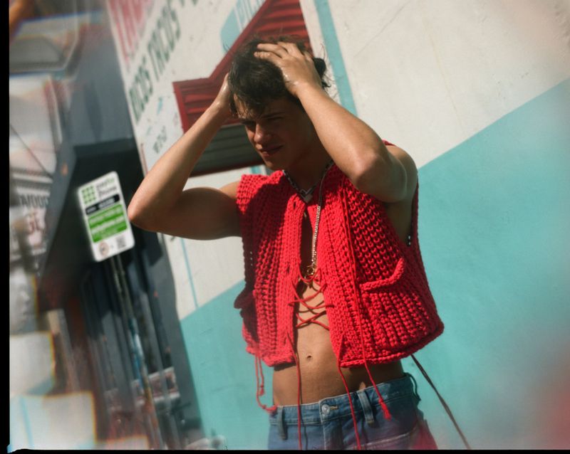 Young adult male in a red knit vest, hand in hair, posing outdoors with a partially visible building with green and white signage.
