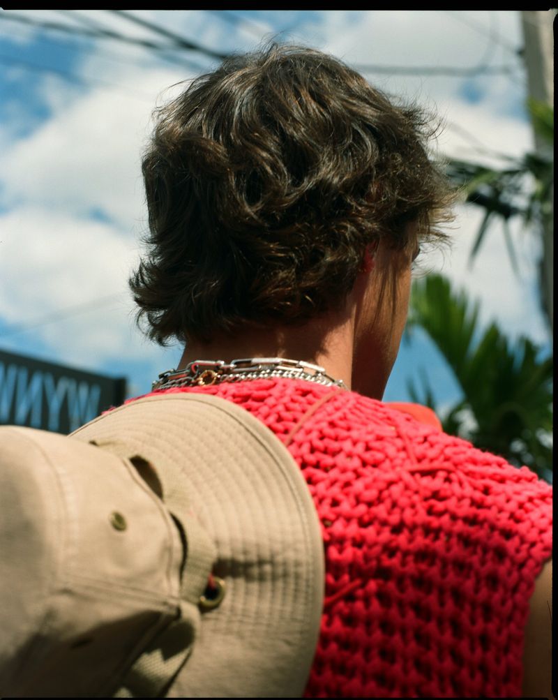 From behind, a person with brown hair and a coral knit top is shown outdoors, wearing a necklace and holding a baseball cap.