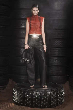 A fashion model poses in a rust-colored top and black pants on a tire, against a tire background in a studio setting with dramatic lighting.