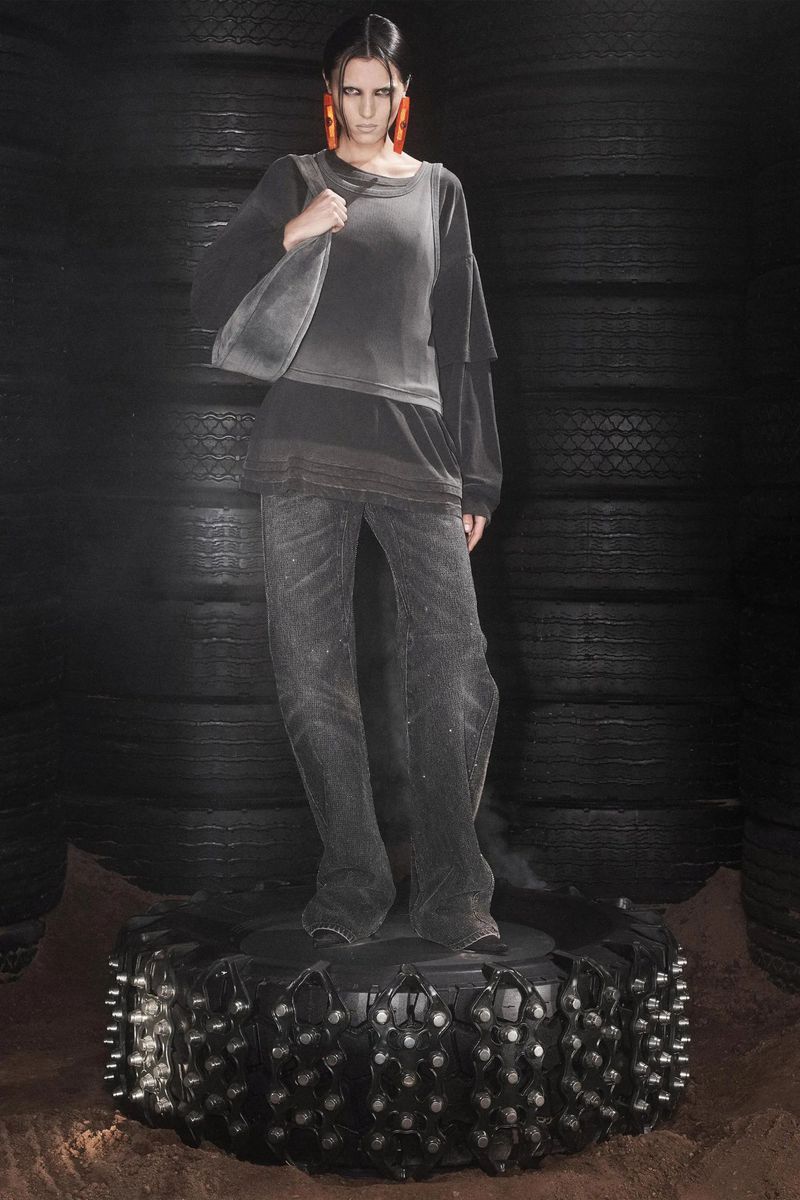 A woman in a black outfit stands on a studded tire, posing in front of a tire wall, lit in dark studio lighting for a fashion shot.
