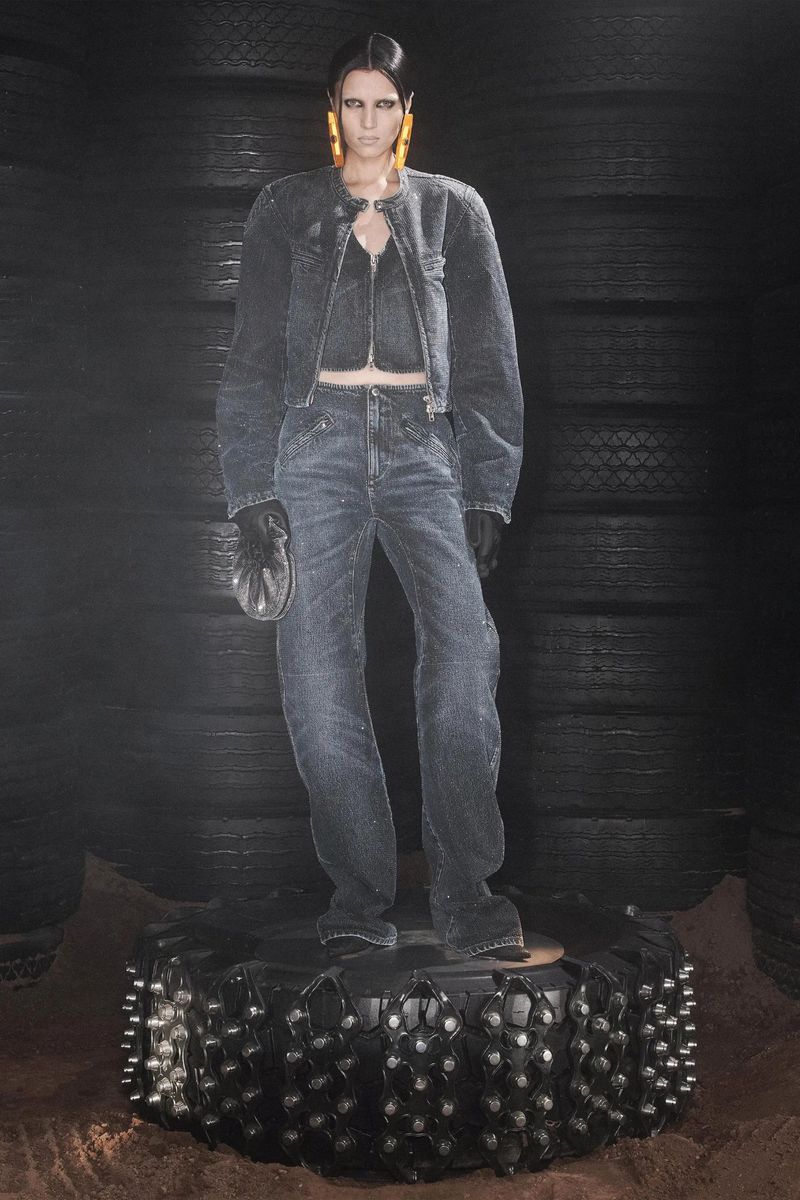 Woman poses in dark denim jacket and jeans on a studded tire pedestal against a backdrop of stacked tires, fashion editorial style.