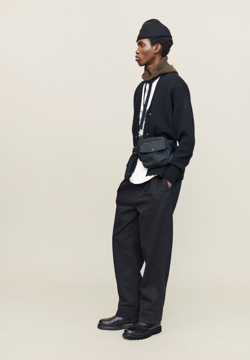 Studio shot of an adult male wearing a black beanie, cardigan, pants, and shoes, standing against an off-white backdrop with a messenger bag.