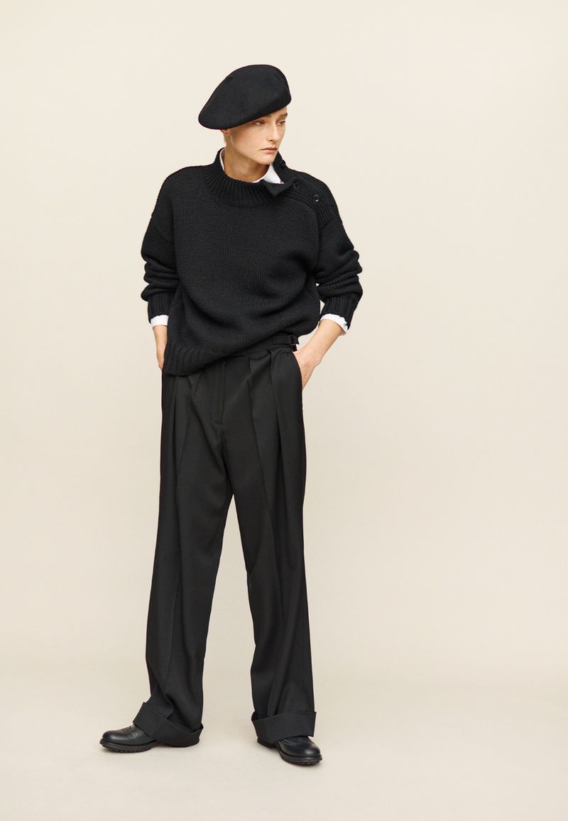 Adult female model in a full-length studio portrait wearing a black sweater, black trousers, black shoes and a black beret, looking to the side.