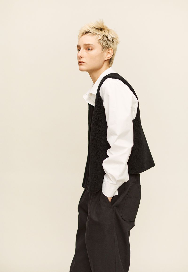 Model with short blonde hair standing in profile wearing a black vest over a white collared shirt and black trousers.