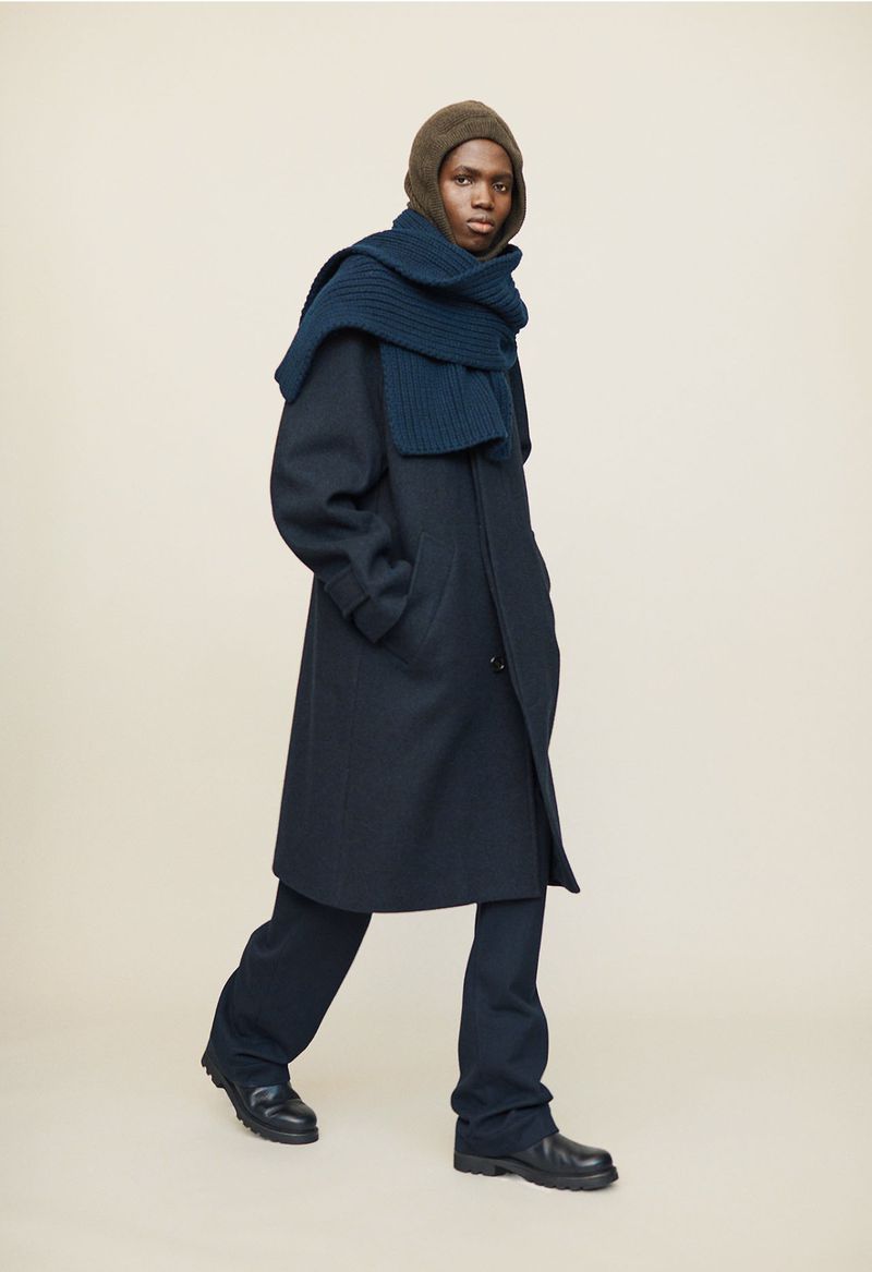 Fashion shot of a Black male model wearing a dark navy overcoat with matching pants, a knit scarf, and a knit balaclava, against a light background.