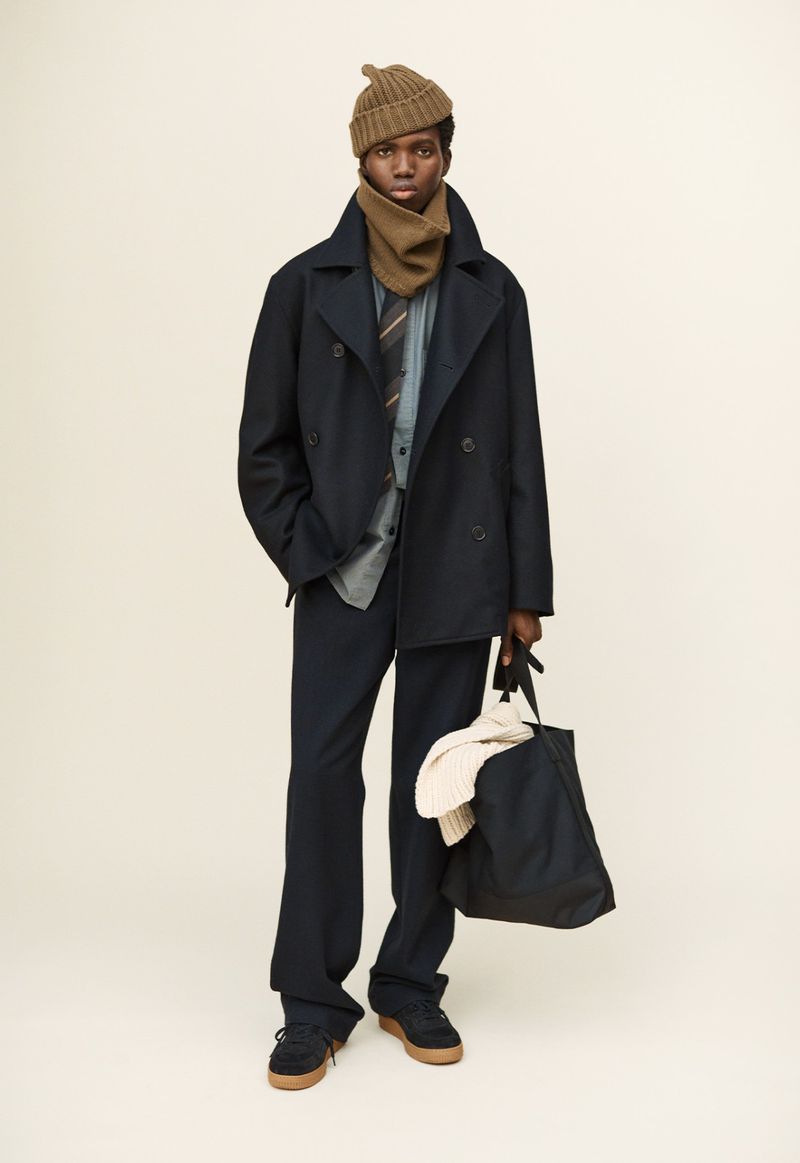 Full-length studio shot of a dark-skinned male model wearing a pea coat, trousers, beanie, scarf, shirt and tie, and holding a tote bag.