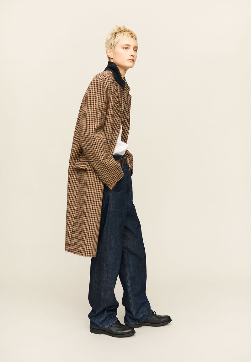 A stylish adult in a brown houndstooth coat, blue jeans, and black shoes poses in a full shot against a neutral studio backdrop.