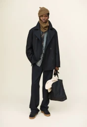 Man wearing a dark double-breasted coat, brown beanie, and neck warmer, holding a large dark tote bag against a neutral studio background.