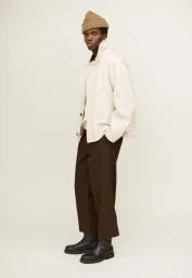 Model in an off-white jacket, dark brown cropped trousers, knit beanie, and black boots posing against a plain backdrop.