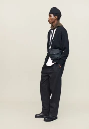 Model wearing a black beanie, cardigan, and trousers with a crossbody bag against a neutral studio backdrop